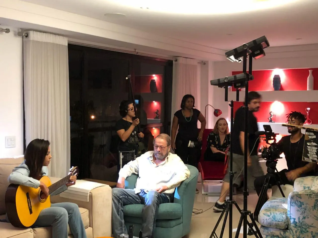 Gravação em interiores: equipa com câmara e iluminação, um músico com guitarra e um entrevistado.