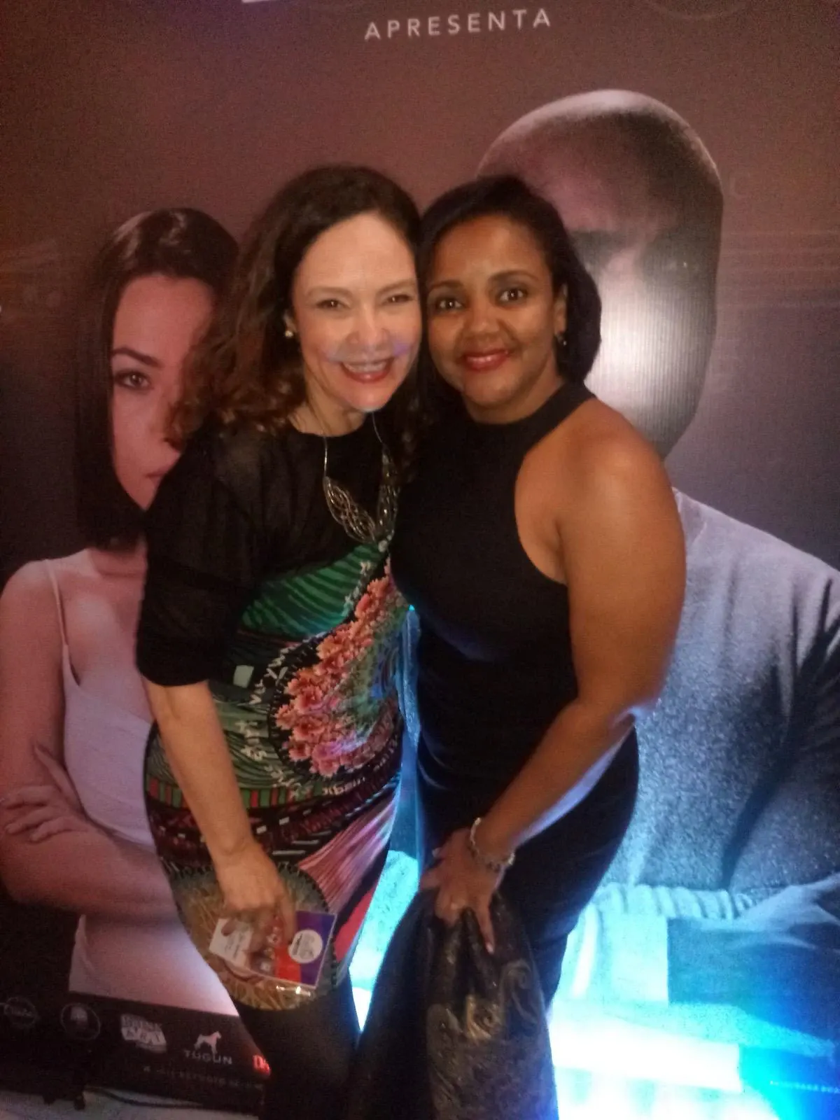 Aline Souza com uma convidada diante de um cartaz de cinema.